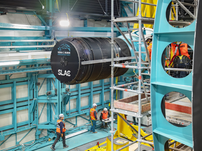 The giant telephoto lens of the LSST Camera as it is being placed on the Rubin Observatory, surrounded by scaffolding. People are below it in orange safety gear.