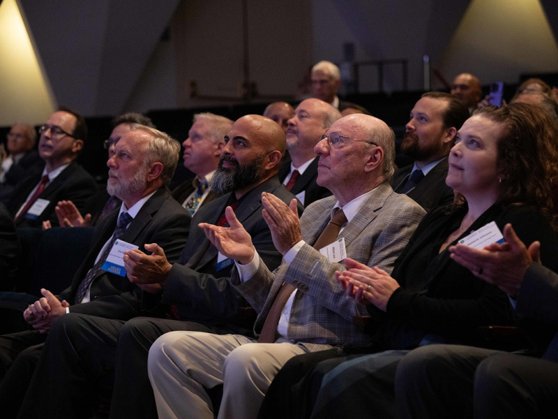 A seated audience clapping while looking at a stage (off screen).
