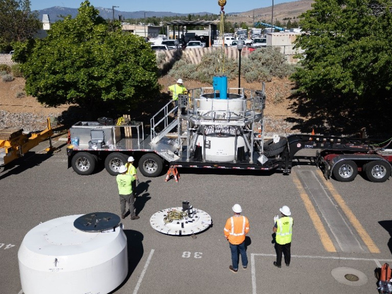 In a parking lot, a crane lowers a blue cylinder into a larger cylinder that is on a large trailer being towed by semi. Four people in high-visibility clothing and white hard hats watch.
