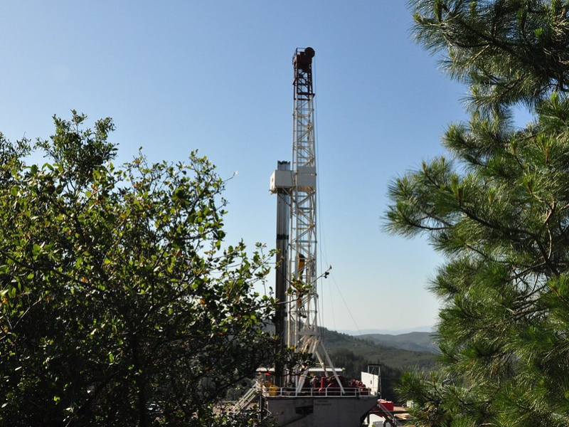 Photo of a drill rig between trees in the foreground, with rolling hills extending into the background.