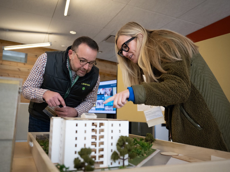 A man and a woman standing near a table that has a Design Challenge team entry house design model sitting on it; they're looking at the details of the model.