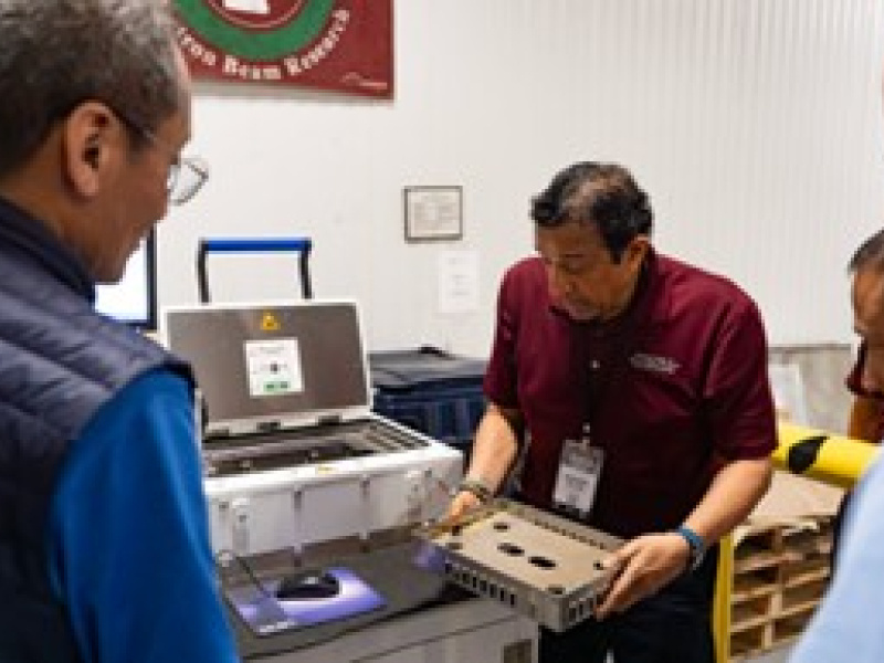 Standing next to a machine, a man holds something looking like a tray. Three people look on.