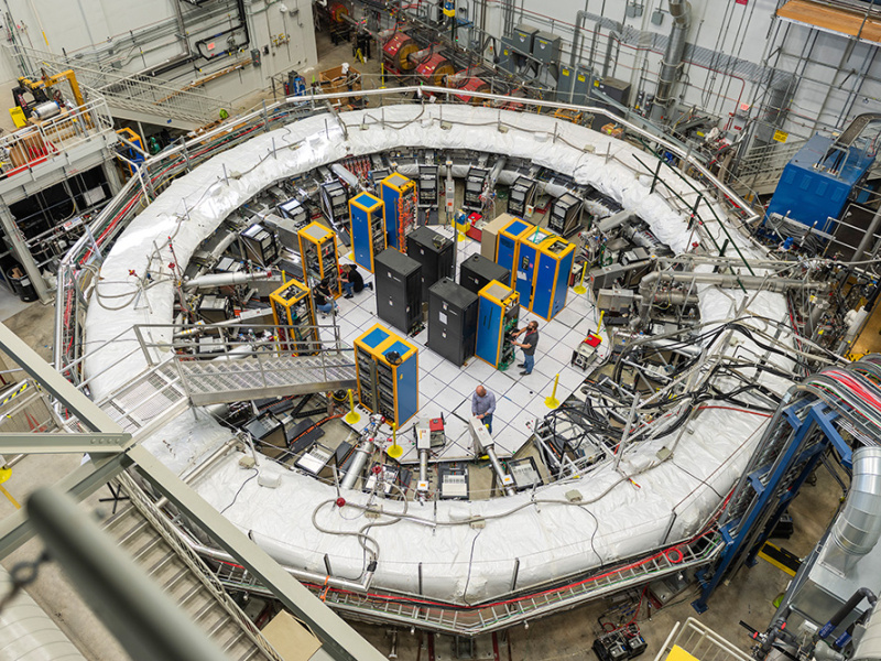 An aerial view of the Muon g-2 experiment, which has a large ring covered in white padding. There are metal arms and tubes coming out of the ring into the middle. There are also computer cabinets and scientists standing inside of the ring. An aerial view of the Muon g-2 experiment, which has a large ring covered in white padding. There are metal arms and tubes coming out of the ring into the middle. There are also computer cabinets and scientists standing inside of the ring. 