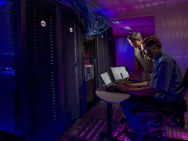 Two men operate a large collection of computers using laptops. 