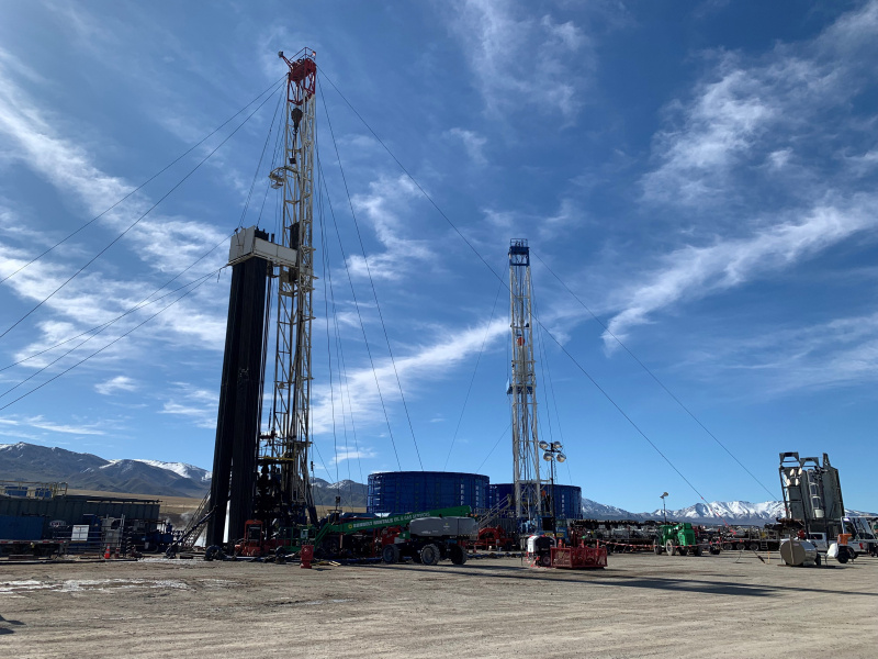 Two drills rigs stretch up into a blue sky with whispy clouds, surrounded by shorter equipment. A snow-capped mountain range is visible in the distance.