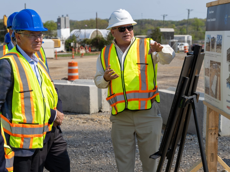 People in reflective vest and hard hats gather around a display and listen to a speaker.