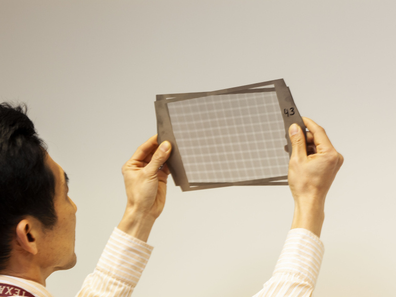 A view over the shoulder of a man holding two overlapping metal sheets over his eye level. The silver metal sheets each have a narrow frame that around a rectangular area perforated with many small holes. The holes in the two sheets line up to create a moire pattern.