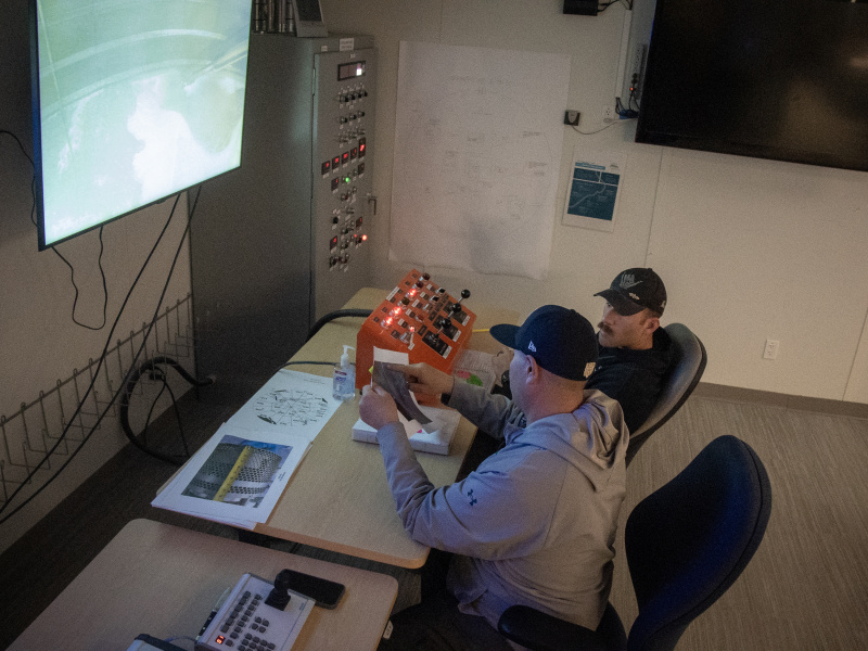 Two men at a control panel desk with a screen on the wall in front of them