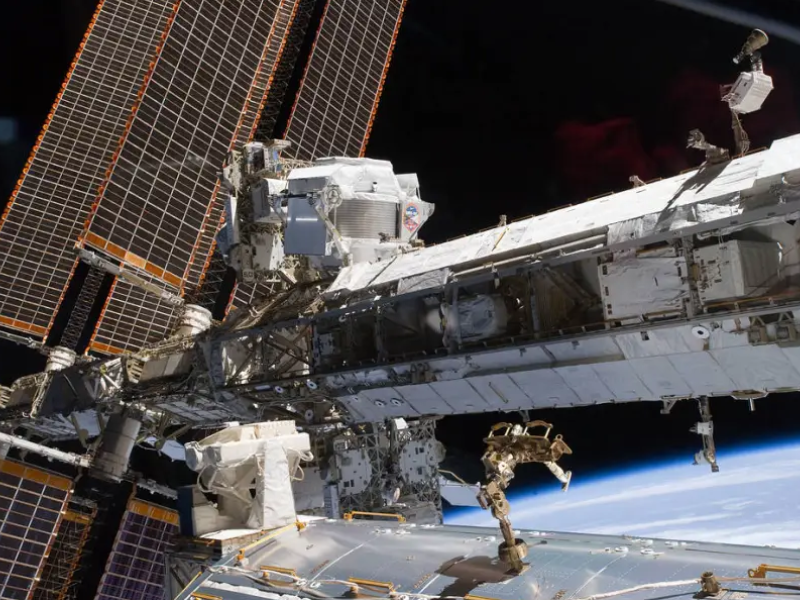 The long tube of the International Space Station runs from left to right across the photo. On the left side, further away from the viewer, large solar panels run from top to bottom. There is a white box-shaped machine just in front of the solar panels, which is the AMS-02. The blackness of space and Earth’s horizon are in the background.