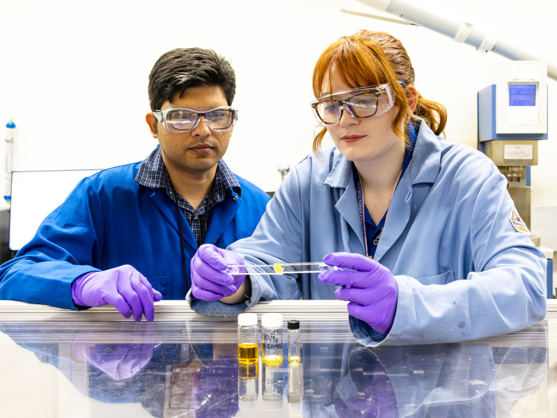 Mary Danielson, who is wearing a lab coat, goggles, and gloves, is holding two pieces of glass attached together with the yellow adhesive. She is leaning on a reflective lab bench with samples of the adhesive in small glass bottles in front of her. Anisur Rahman (who is also in lab equipment) is standing next to her, watching. 
