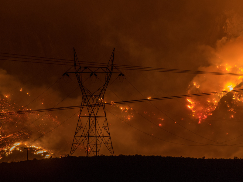 Transmission towers and power lines silhouetted against a large wildfire burning across a hillside at night.