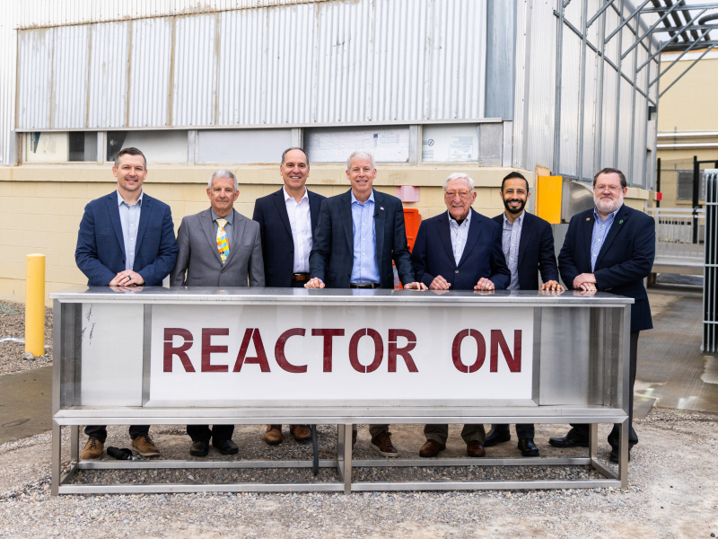 A group photo of DOE leadership standing in front of a sign that says "Reactor On" 