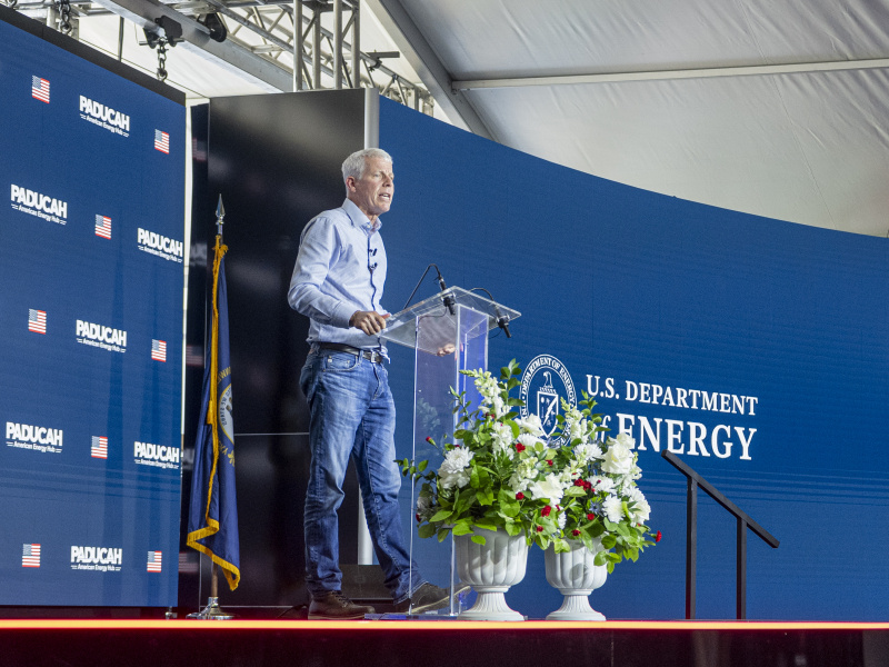 The US Department of Energy Secretary Chris Wright standing on a stage behind a clear podium with a blue backdrop behind him with the words Paducah Site and US Department of Energy written on the backdrops