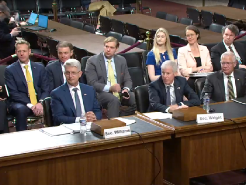 Three men in business suits look to the left of the photo. Behind them are a row of six people also looking to the left. From left, name tags say Sec. Williams., Sec. Wright, and Sec. Walsh.