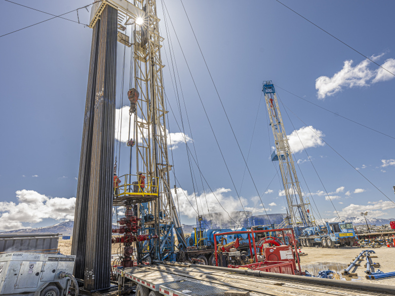 Close-up photo of a drill rig stretching up into a blue sky with the sun shining right behind the derrick (i.e. the tower).