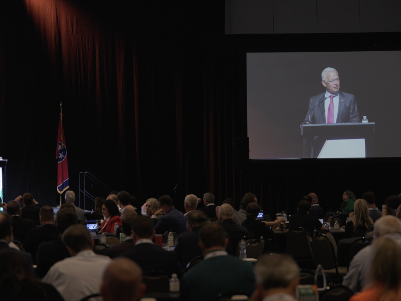 EM Assistant Secretary giving a speech, on a stage standing behind a podium, to a large crowd. To the right he is displayed on a large screen