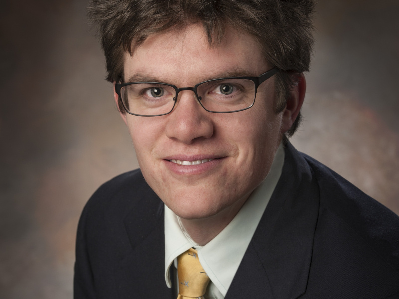 Headshot of a white man with brown hair and glasses in a black suit with gold tie