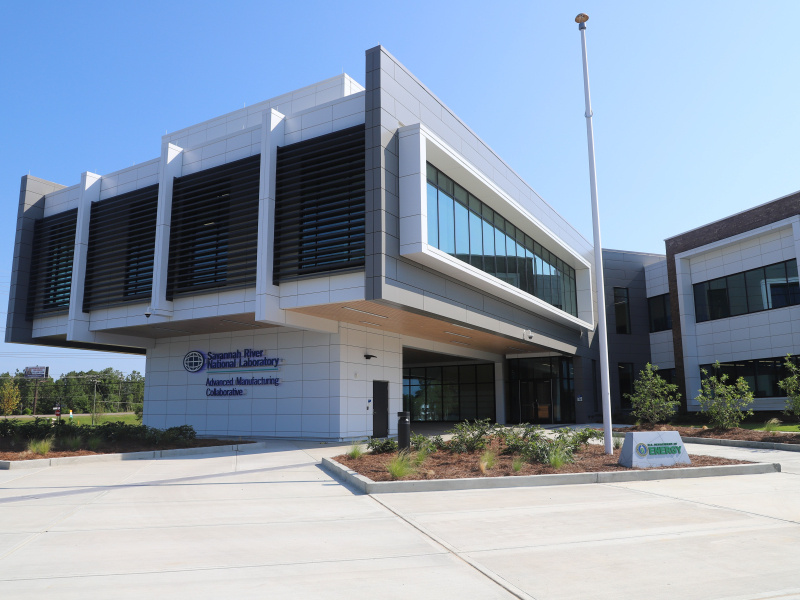 A large white building with a large overlook area in the front with lots of windows. There are plants around the building and trees in the background