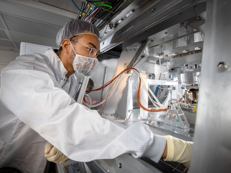 A man in a clean room suit is looking at a complex piece of machinery within a metal cabinet.