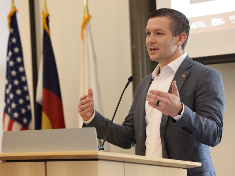 A man speaks at a lectern with American flags and the corner of a powerpoint on a large display screen in the background.
