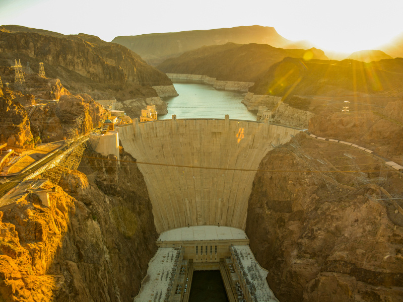 The sun rises over the hoover dam with the memorial bridge and reservoir visible in the shot