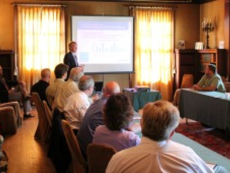 Group of people sitting at a table in a room, with a man speaking at the front and a display screen behind him.