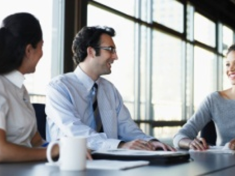 Three people in business attire sitting at a table talking / laughing.