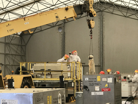 Rigging and heavy equipment operators guide a crane during the removal of stainless-steel cylinders containing tritium gold traps from a concrete culvert.