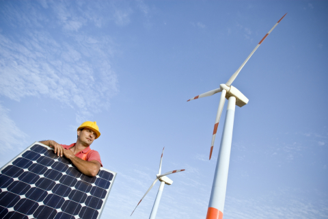 A male construction worker stands by a solar panel with a wind turbine in the background. 