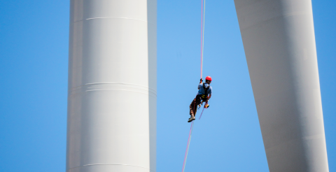 Worker hanging from cable between wind turbine rotor and tower with blue sky behind him.