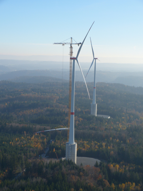 During construction, workers climb out on top of a tall wind turbine and guide 19-ton turbine blades into place.