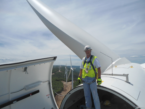 Man in a harness standing on top of an open wind turbine with other wind turbines in the distance.