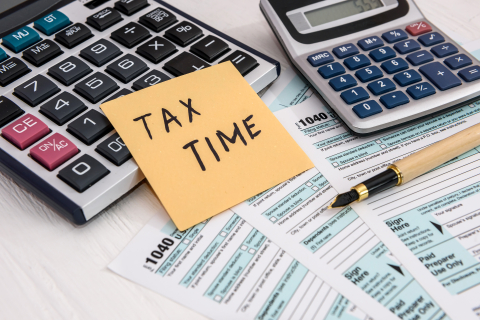 An orange post-it note that says "tax time" is laid across a calculator. Another calculator is beside the first calculator on top of tax return forms and next to a pen.