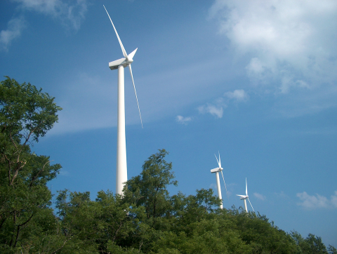 Three wind turbines behind a green tree line and against a blue sky with light, wispy clouds.