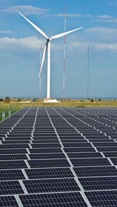 A large wind turbine stands against a blue sky, towering over a field of solar photovoltaic panels.