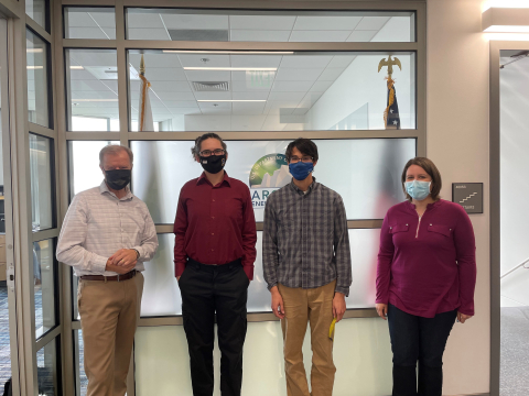 AEO Director George Roe stands to the left of three interns in front of the office wall. All three are wearing masks. 
