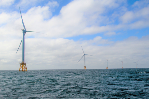 Offshore wind turbines standing tall over the water, against a blue sky with white clouds.