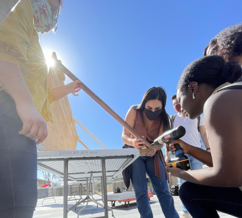 Three women build a wooden structure on a table outside as the sun shines.