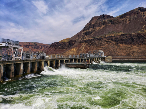 A dam creates churning waters in a river flowing between red hills.