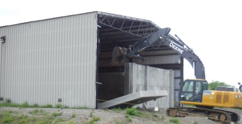 An operator at the West Valley Demonstration Project uses an excavator to remove a large concrete shield wall to create an entrance to accommodate a large fork truck for transporting empty waste containers in and out of the building.