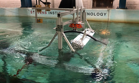 Two metal devices face each other from opposite ends of a wave tank that looks like an indoor swimming pool. Stevens Institute of Technology is written on the inside of the tank wall just above the water surface. Hanging over the water, a network of blue metal beams support a pair of electronic boxes on a track.
