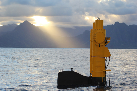 A marine energy device floats on the surface of the ocean. Mountains and cloudy skies are visible in the background. The sun shines through a break in the clouds.