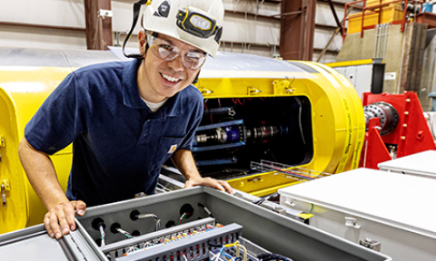 : A researcher in a hard hat leans over a hatch containing electronics and wires.