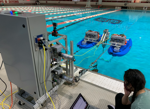 A photo of a researcher sitting next to a pool with a computer while two blue floating structures holding electronics pull the kite through the water.