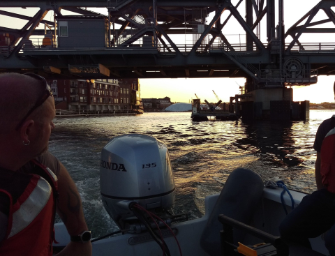 A person driving a motorboat in a narrow channel near a metal bridge at dusk.