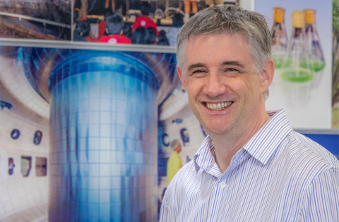 Richard Buttery, Director of the DIII-D National Fusion Facility, stands in front of an image showing the interior of the DIII-D tokamak.