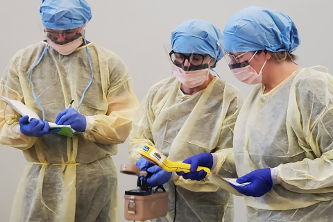 People dressed in heavy-duty medical garb hold and examine a hand-held Geiger counter