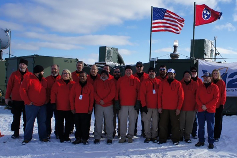Twenty or so people stand outside in the snow wearing matching red jackets. A U.S. and Tennessee flag fly in the background.