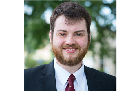 Headshot of a young man with trees in the background. He is wearing a black suit with a white shirt and red tie and smiling at the camera.