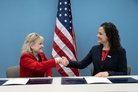 Two women shaking hands after signing a document in front of the American flag.